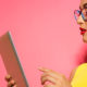 Fashionable lady in yellow in front of a pink background reading about IT marketing on a green tablet.